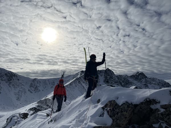 Deux skieurs encordés avec baudriers, leurs skis de randonnée fixés aux sacs à dos, grimpant vers le Pic de Pedrons (2715 m) à travers un terrain mixte de neige et de roches le long de l'arête. Sortie encadré par un guide de haute montagne en Andorre.